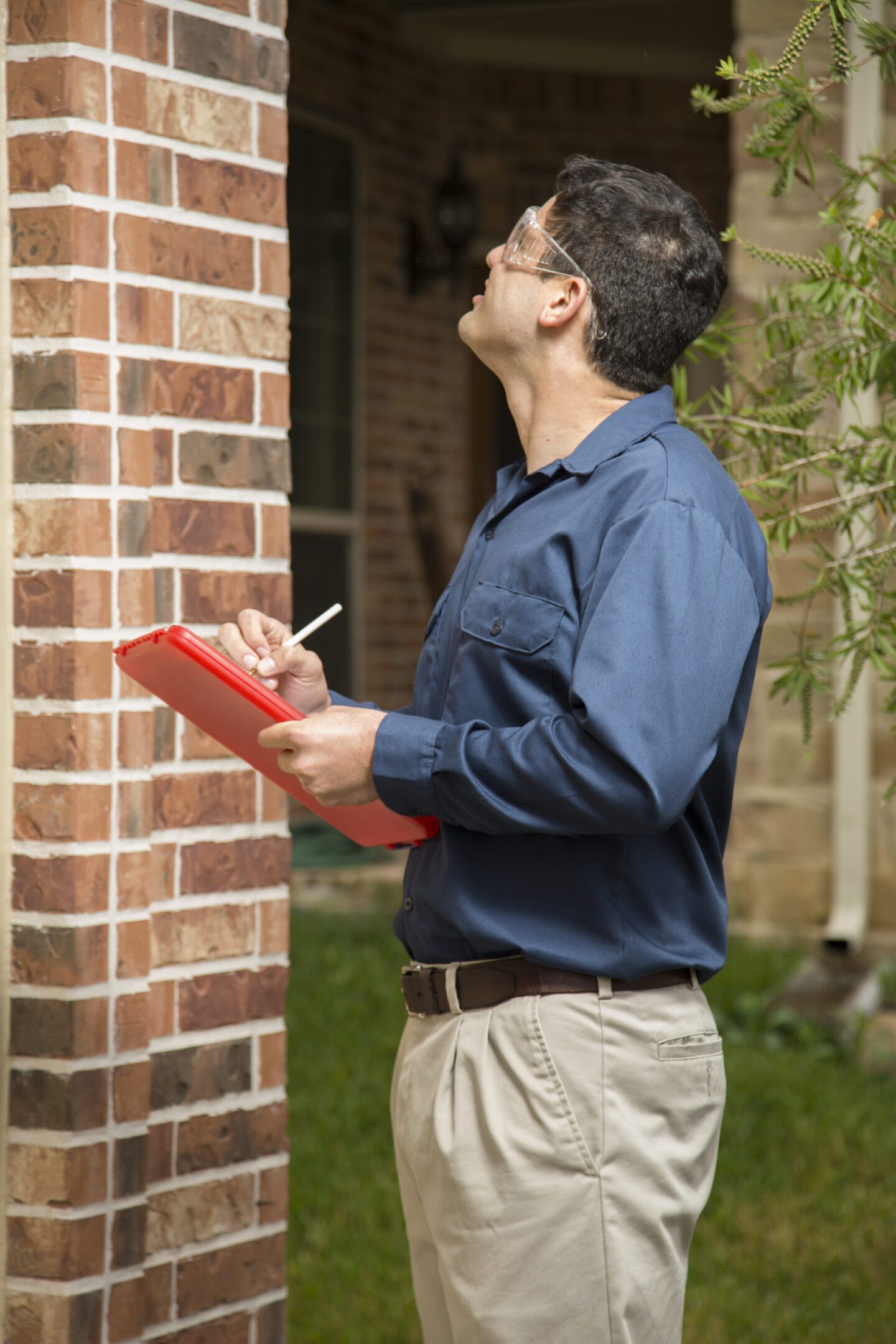 Latin descent repairman, insurance adjuster, or quality control inspector takes notes on clipboard outside red brick home. He is wearing safety glasses and holds a clipboard, pen to write on his report.  Navy and khaki uniform.